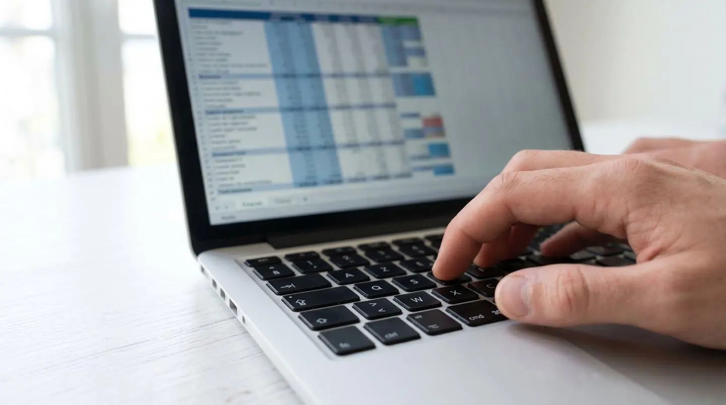 Gros plan sur une main tapant sur le clavier d'un ordinateur portable moderne, feuille de calcul financier partiellement visible et floutée à l'écran, bureau blanc minimaliste